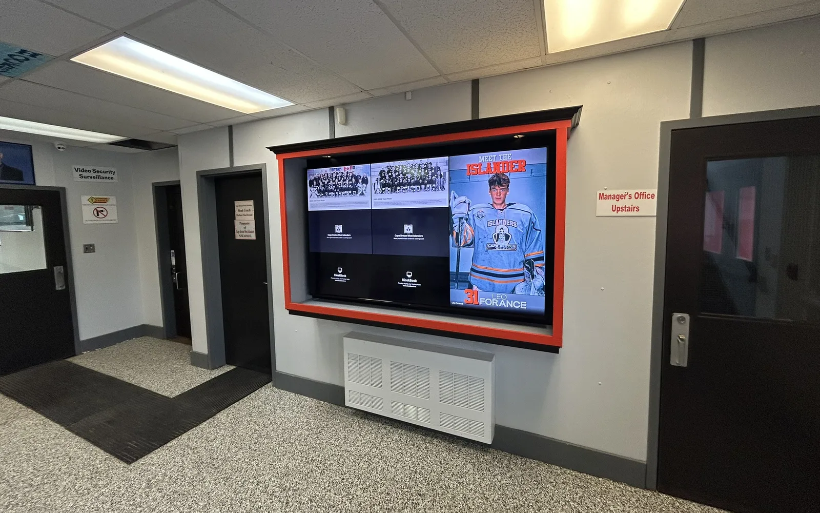 A rink lobby with a large wall-mounted TV in an orange frame. KioskBook shows the home team’s past team photos beside a “Meet the Islander” player profile.